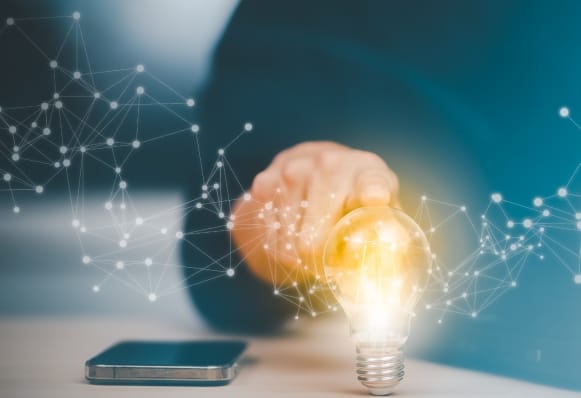 スマートフォンの隣で光る電球に触れる指とデジタルネットワークの表現